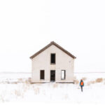 Photo of person standing in snow near a white house with an open window